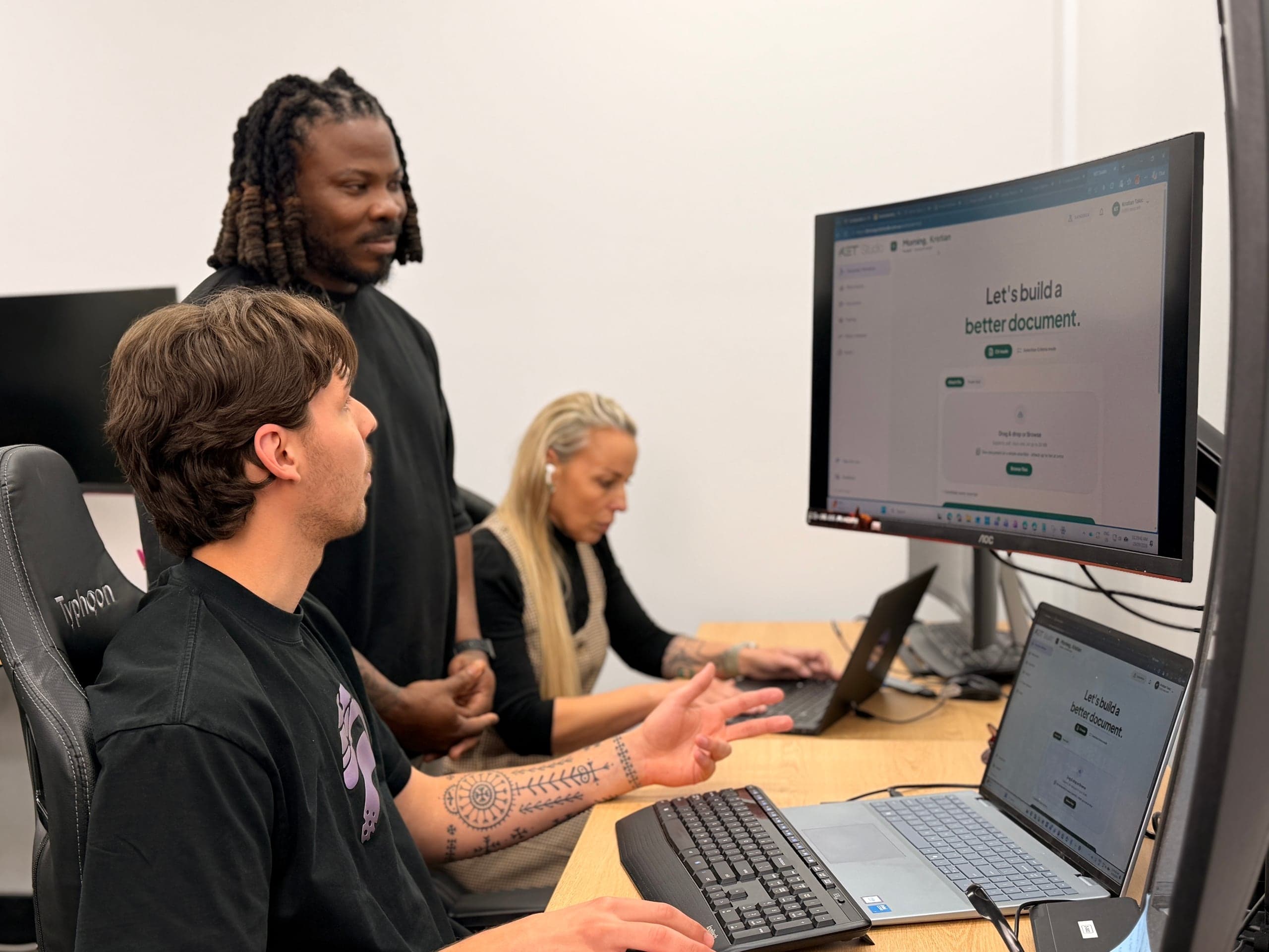 Three colleagues collaborate at a desk, with a man standing beside two seated teammates.